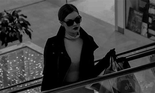 Zu sehen ist ein schwarz weiß Bild auf dem eine chic gekleidete Frau mit Sonnenbrille und dunklem Lippenstift in einem Einkaufszentrum die Rolltreppe hochfährt und einige Einkaufstüten in beiden Händen hält.